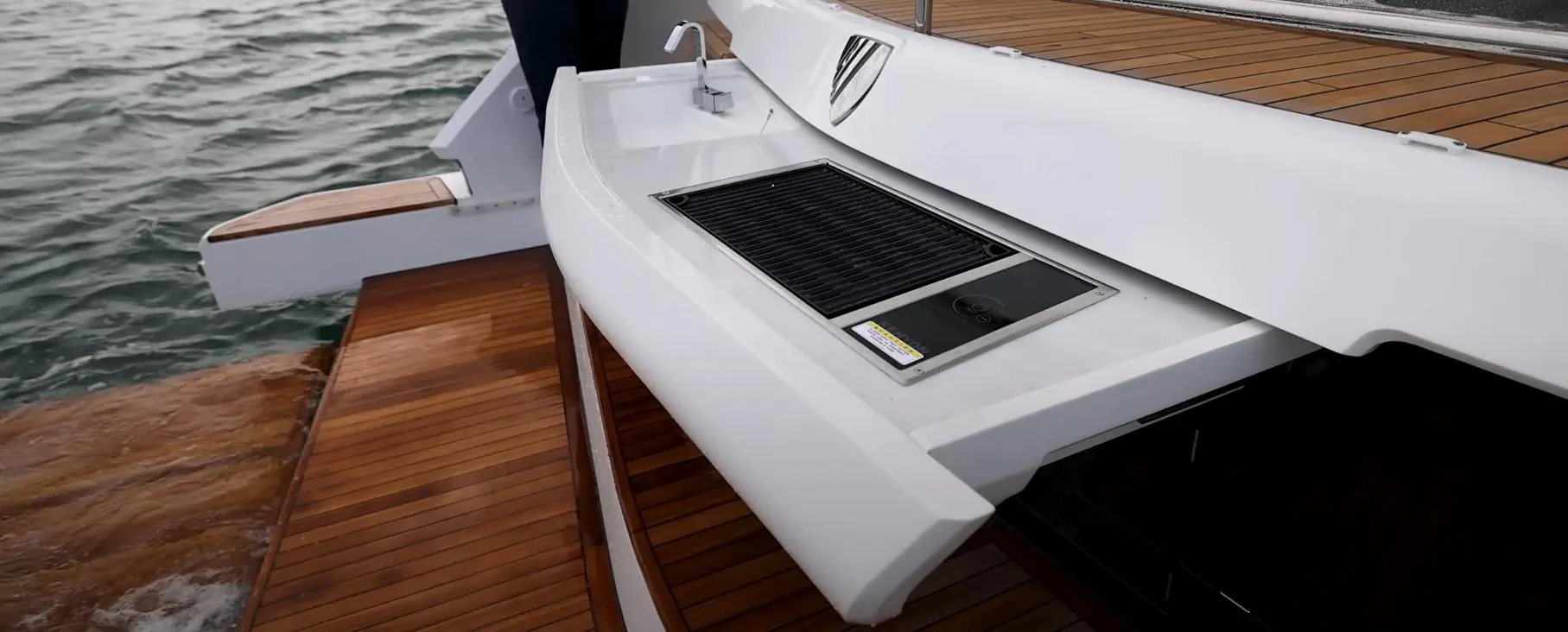 A compact boat kitchen features a stainless steel grill and sink, set against a wooden deck and water backdrop.